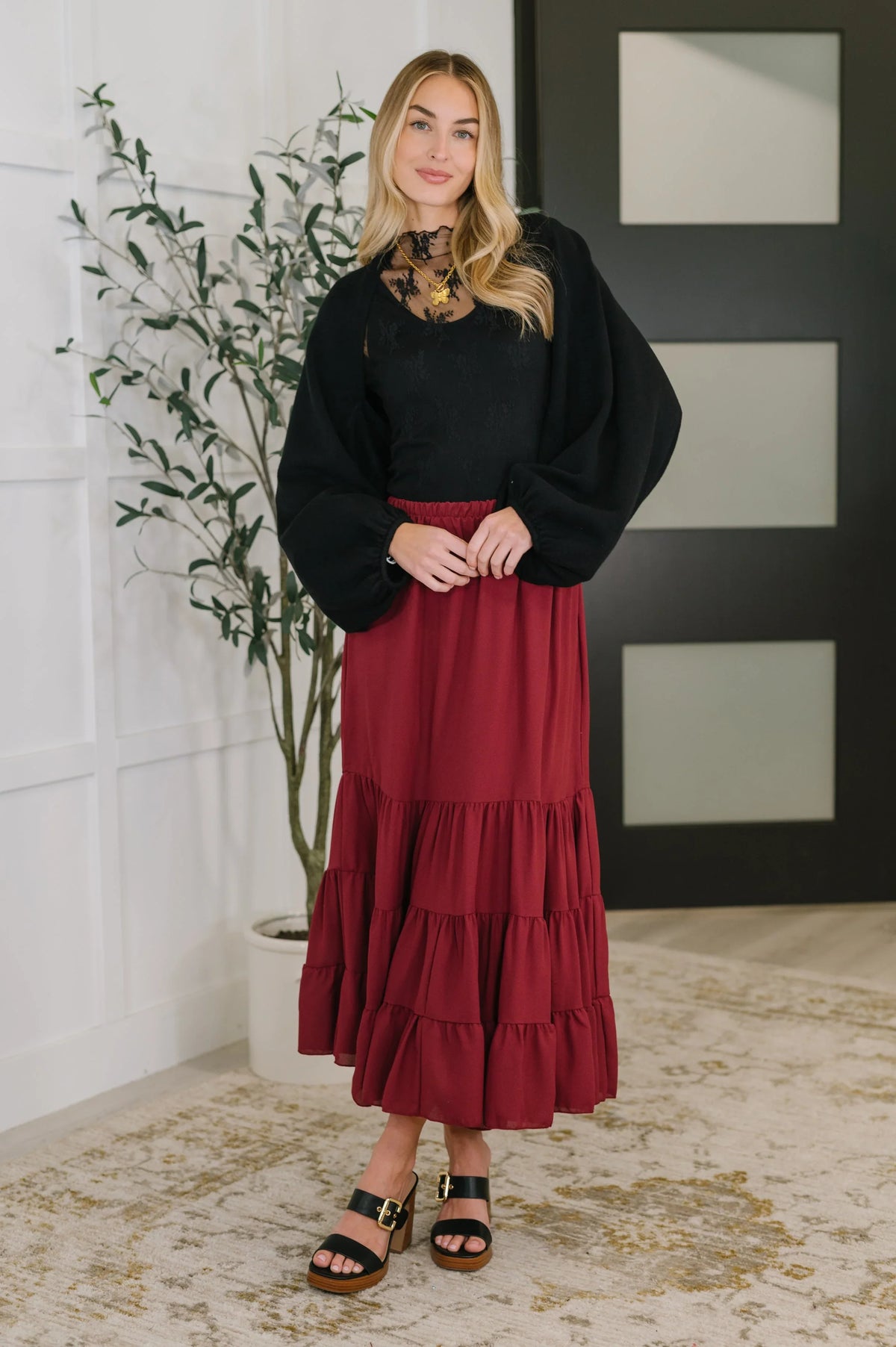 Woman wearing a tiered burgundy maxi skirt with a black lace top and heeled sandals indoors.