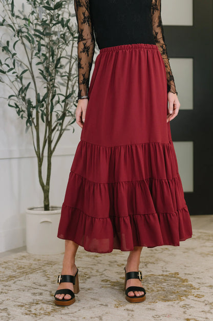 Woman wearing tiered burgundy maxi skirt with black lace top and block heel sandals indoors