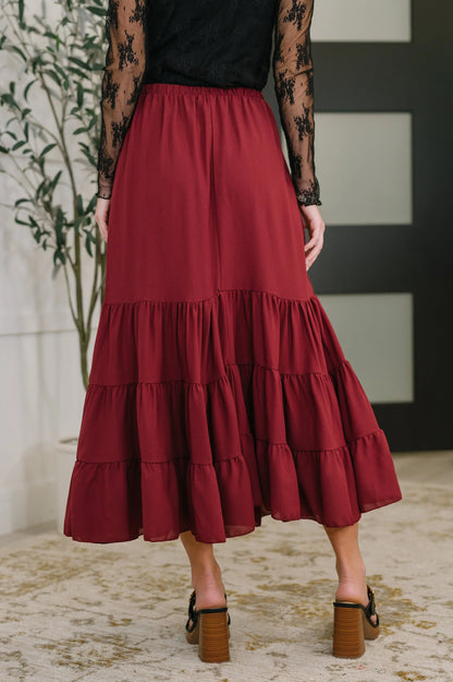 Woman wearing a burgundy tiered maxi skirt with ruffle details and block heel sandals indoors.