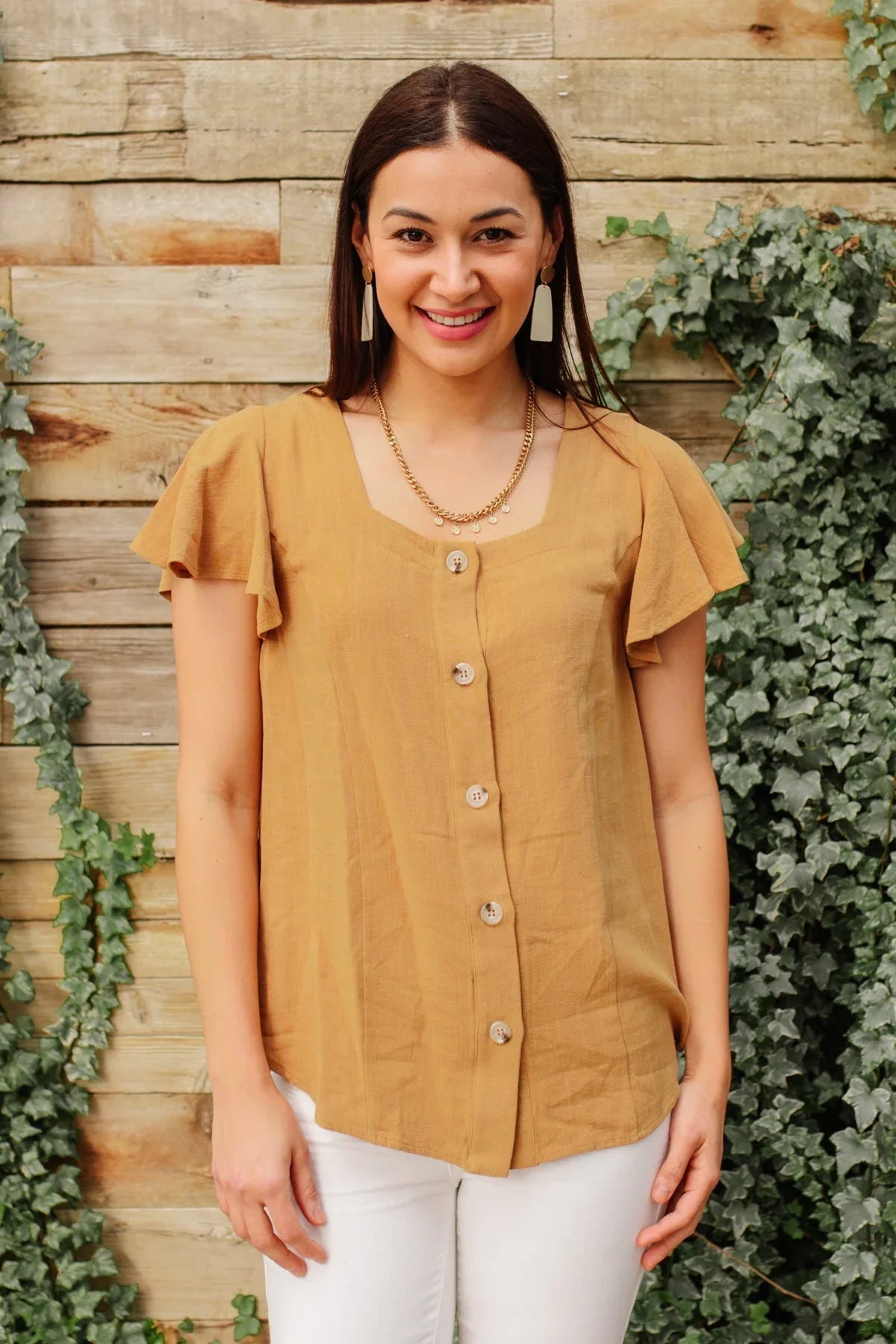 Woman wearing a taupe button-front flutter sleeve blouse with white pants, outdoor setting