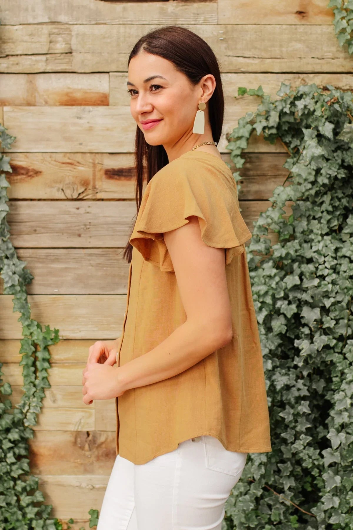 Woman wearing a taupe flutter sleeve top and white pants, standing by a wooden wall with ivy.