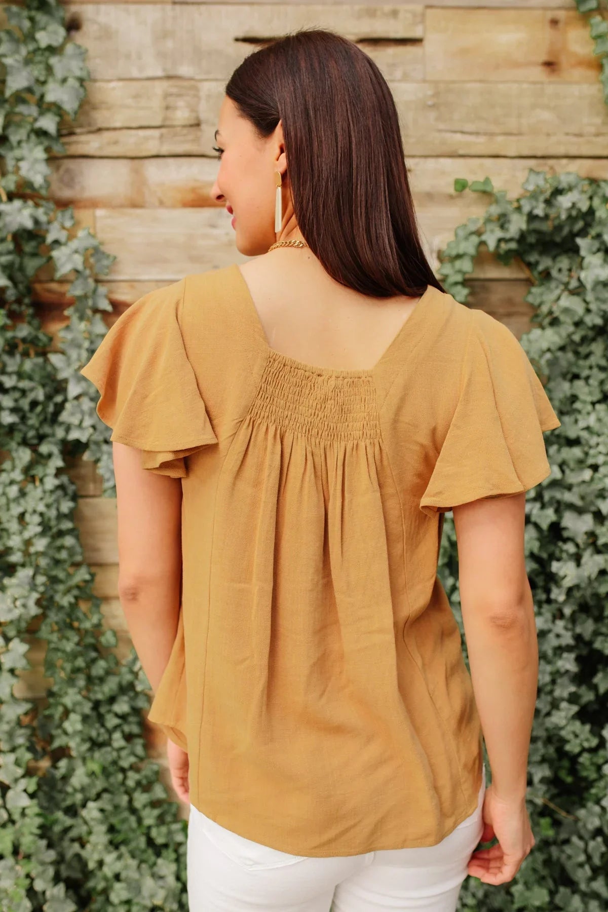 Woman wearing a taupe flutter sleeve blouse with smocked back, paired with white pants.
