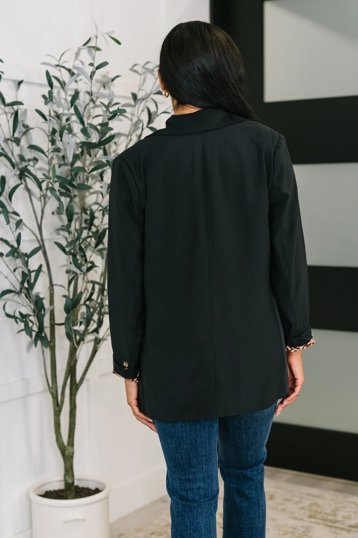 Woman wearing a black everyday blazer and blue jeans, viewed from the back indoors.