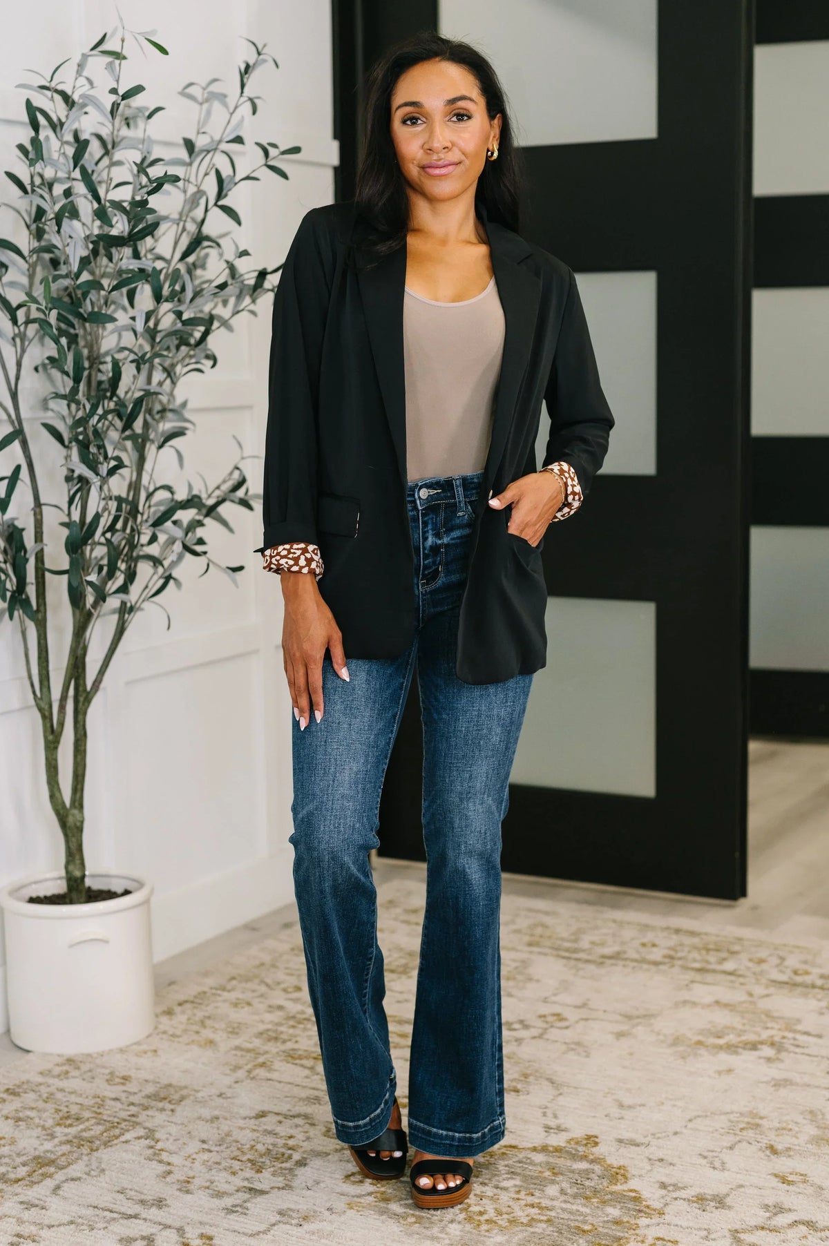 Woman wearing black blazer with patterned cuffs, beige top, and blue jeans in modern room