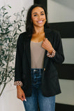 Woman modeling black relaxed-fit blazer with patterned cuffs, taupe top, and blue jeans indoors.