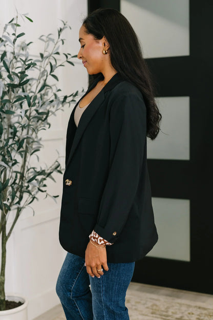 Woman wearing a black blazer with gold buttons, beige top, and blue jeans indoors