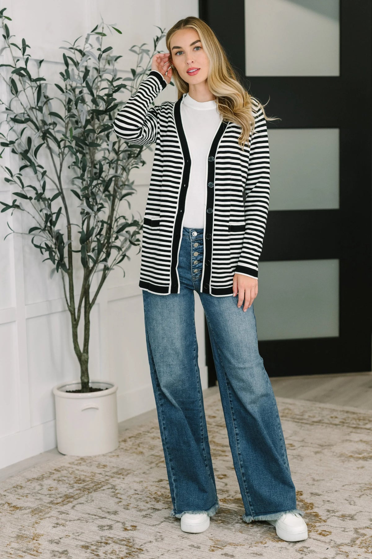 Woman wearing a black and white striped cardigan with jeans and white sneakers indoors.