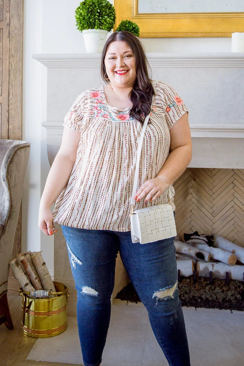 Woman wearing a floral embroidered blouse and distressed jeans with a white crossbody bag indoors