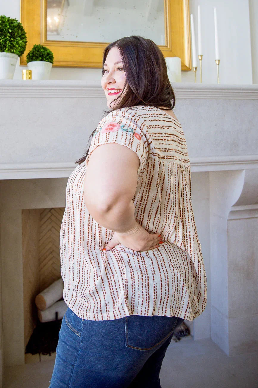 Woman wearing a short sleeve cream blouse with dotted stripes and floral accents, paired with blue jeans