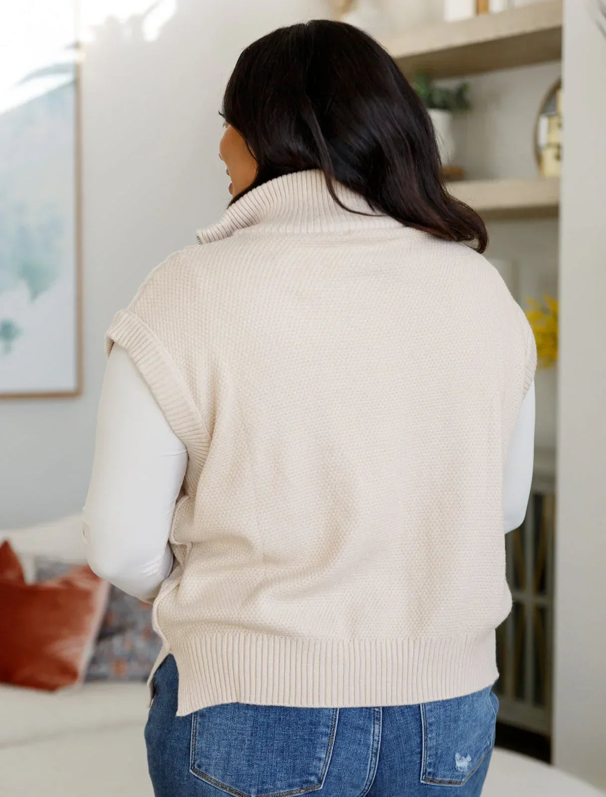 Woman wearing a cream cable knit sleeveless sweater vest with jeans, shown from behind.