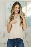 Woman wearing a cream cable knit zip sweater vest and blue jeans in a modern living room.