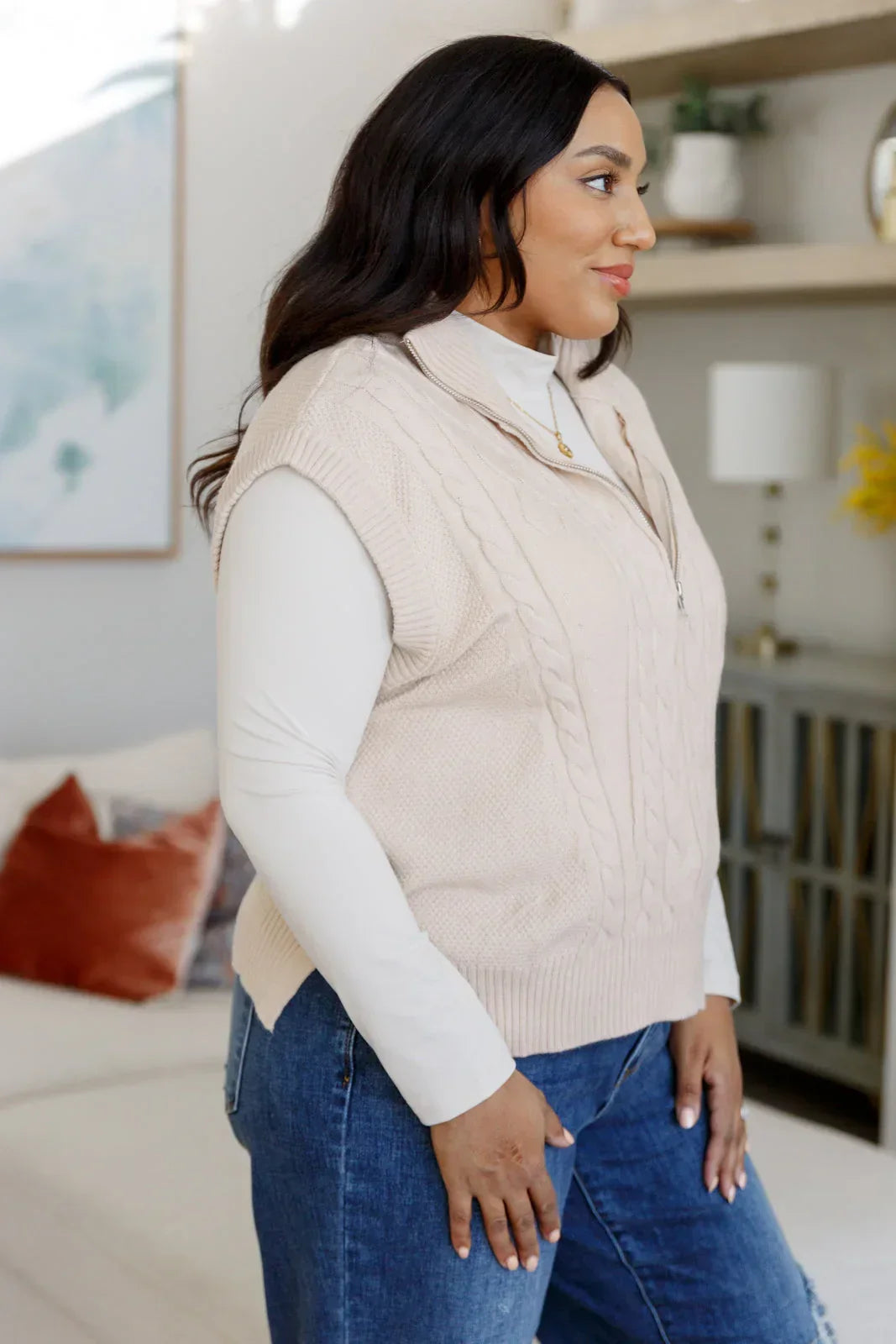 Woman in a beige cable knit zip vest layered over a white turtleneck and blue jeans indoors