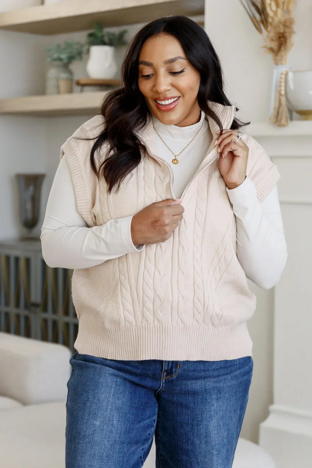 Woman wearing a cream cable knit zip collar vest over a white long sleeve top and blue jeans.