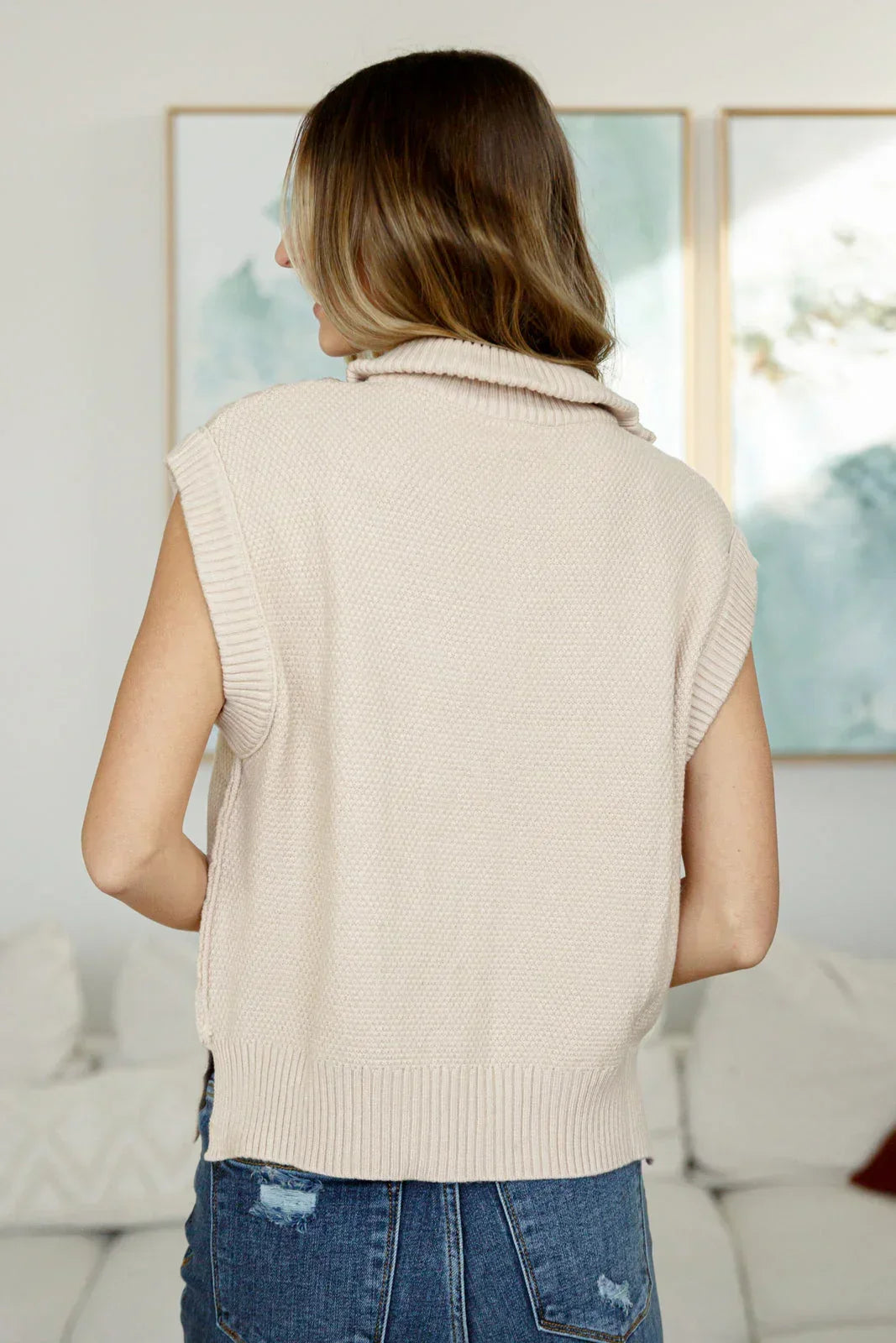 Back view of woman wearing a sleeveless beige cable knit sweater vest and blue jeans indoors