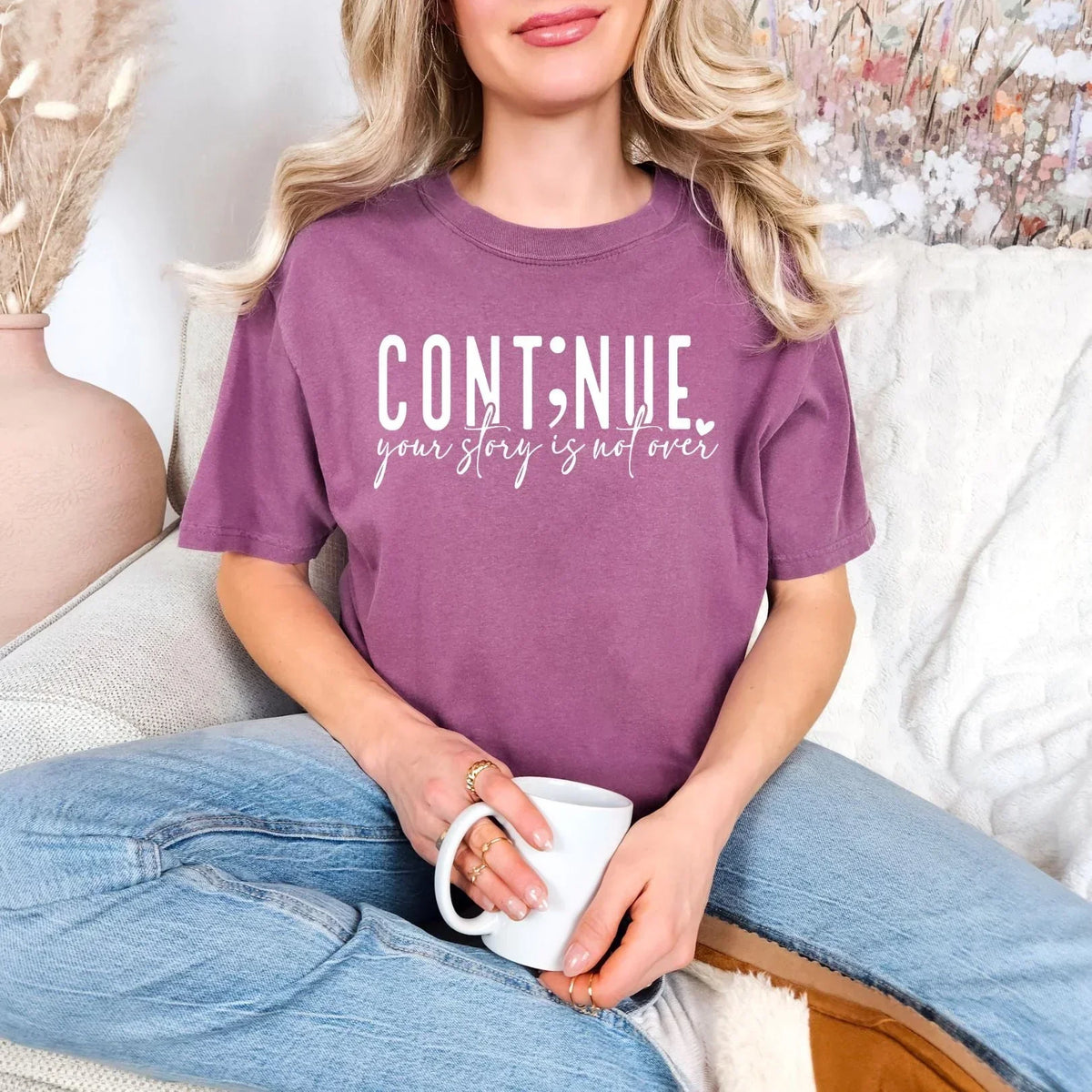 Woman in a purple graphic tee with 'Continue your story is not over' text, sitting casually with a coffee mug.