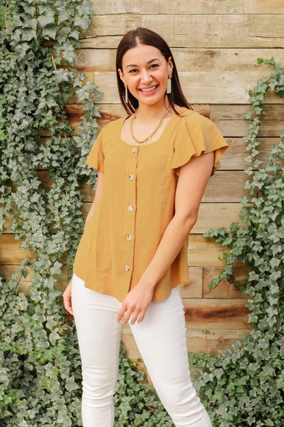 Woman wearing a mustard short-sleeve button front blouse and white pants outdoors