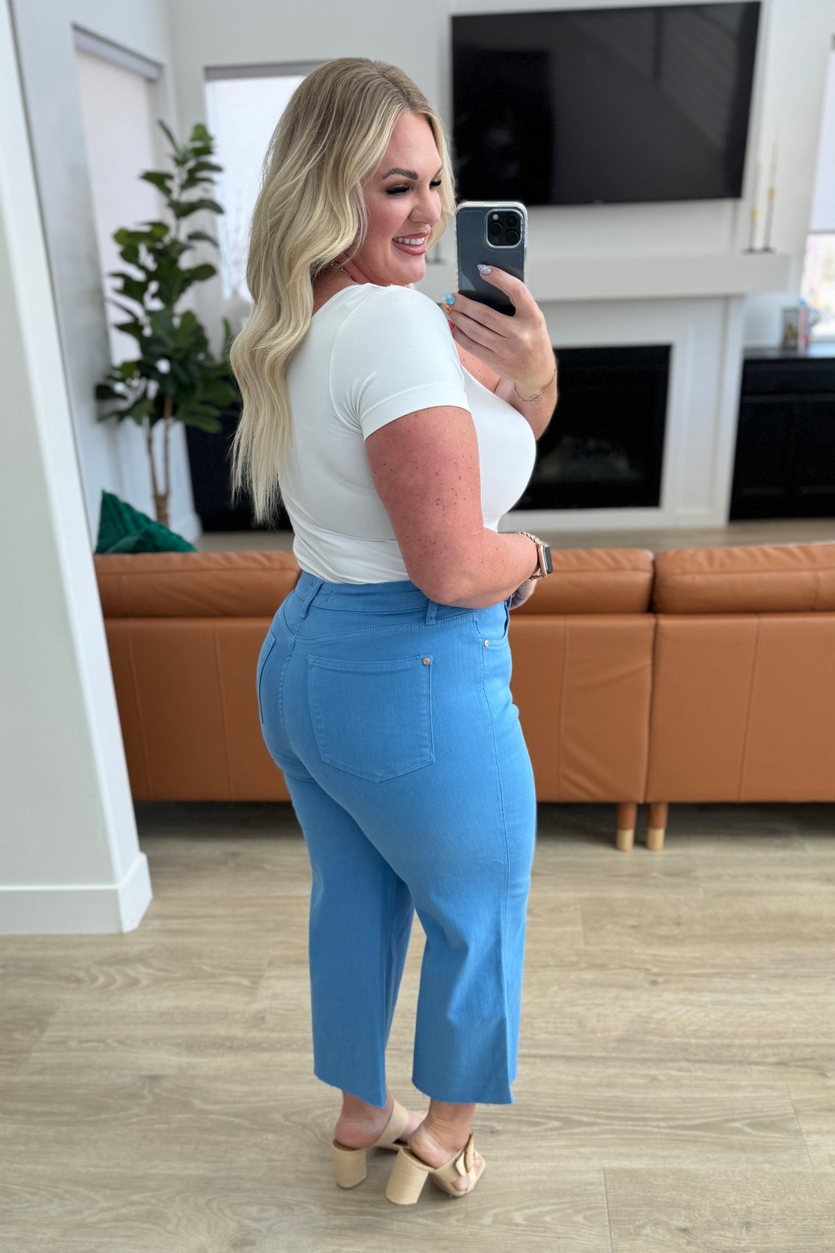 Woman taking a mirror selfie wearing a white top and blue pants in a living room.