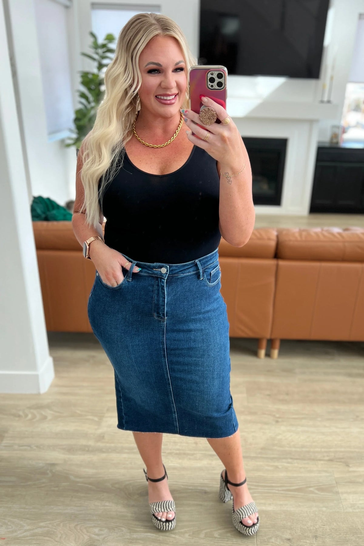 Woman wearing a black tank top with a blue denim midi skirt and platform sandals, posing indoors
