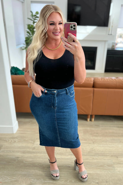 Woman wearing a black tank top with a blue denim midi skirt and platform sandals, posing indoors
