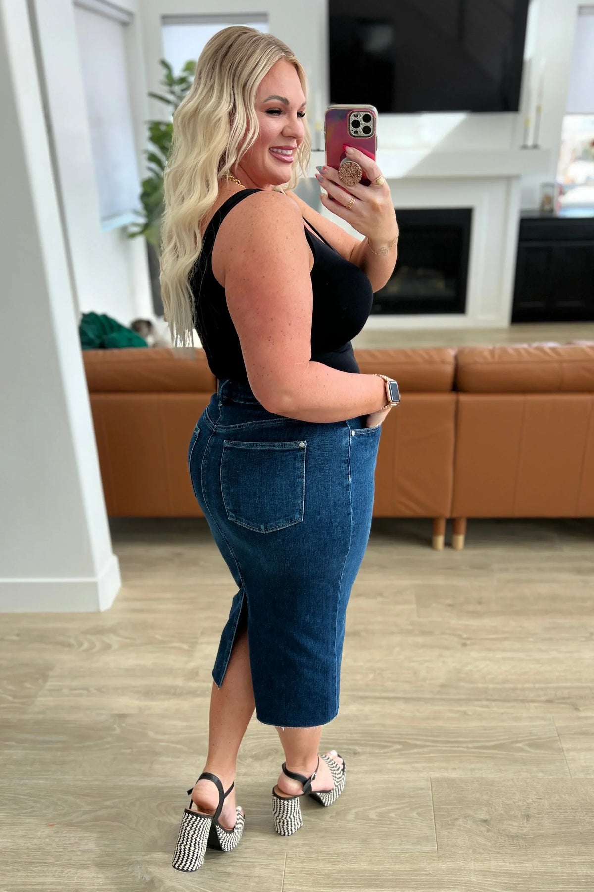 Woman in black tank top and blue denim midi skirt with back slit, wearing heeled sandals, taking a mirror selfie in a modern living room.