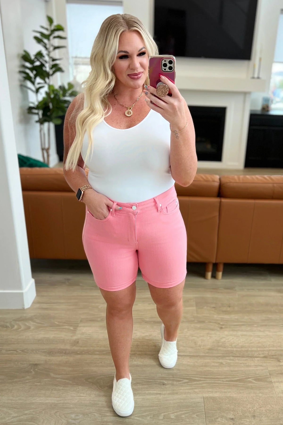 Woman wearing a white tank top and pink denim shorts taking a mirror selfie in a modern living room