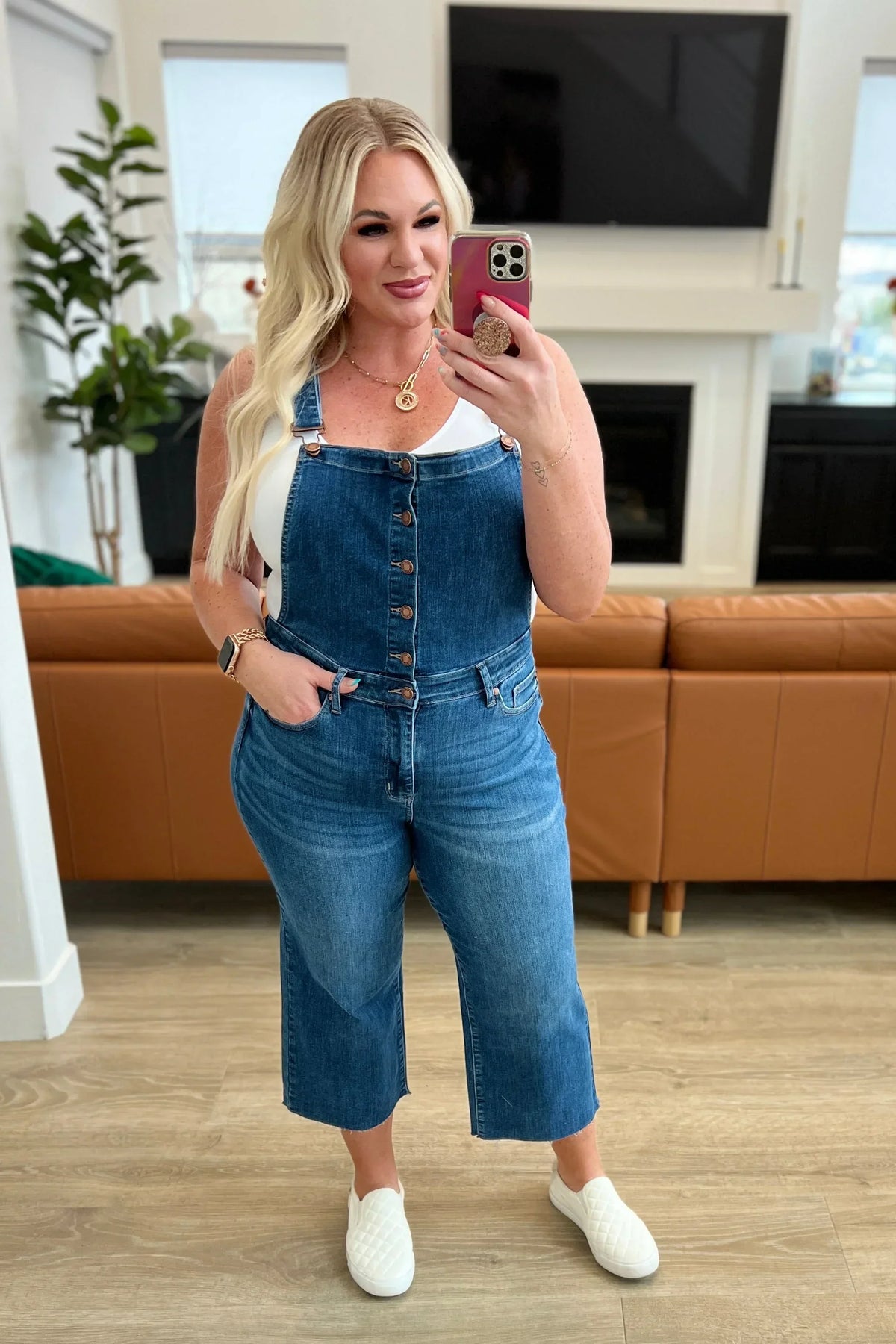 Woman in casual denim overalls and white tank top taking a mirror selfie in a modern living room