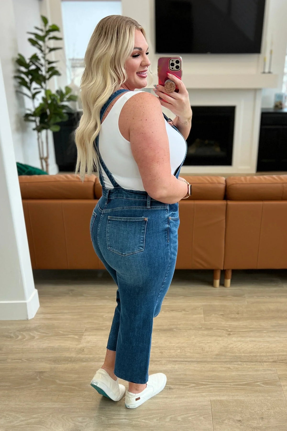Woman in denim overalls and white tank top taking a mirror selfie in modern living room