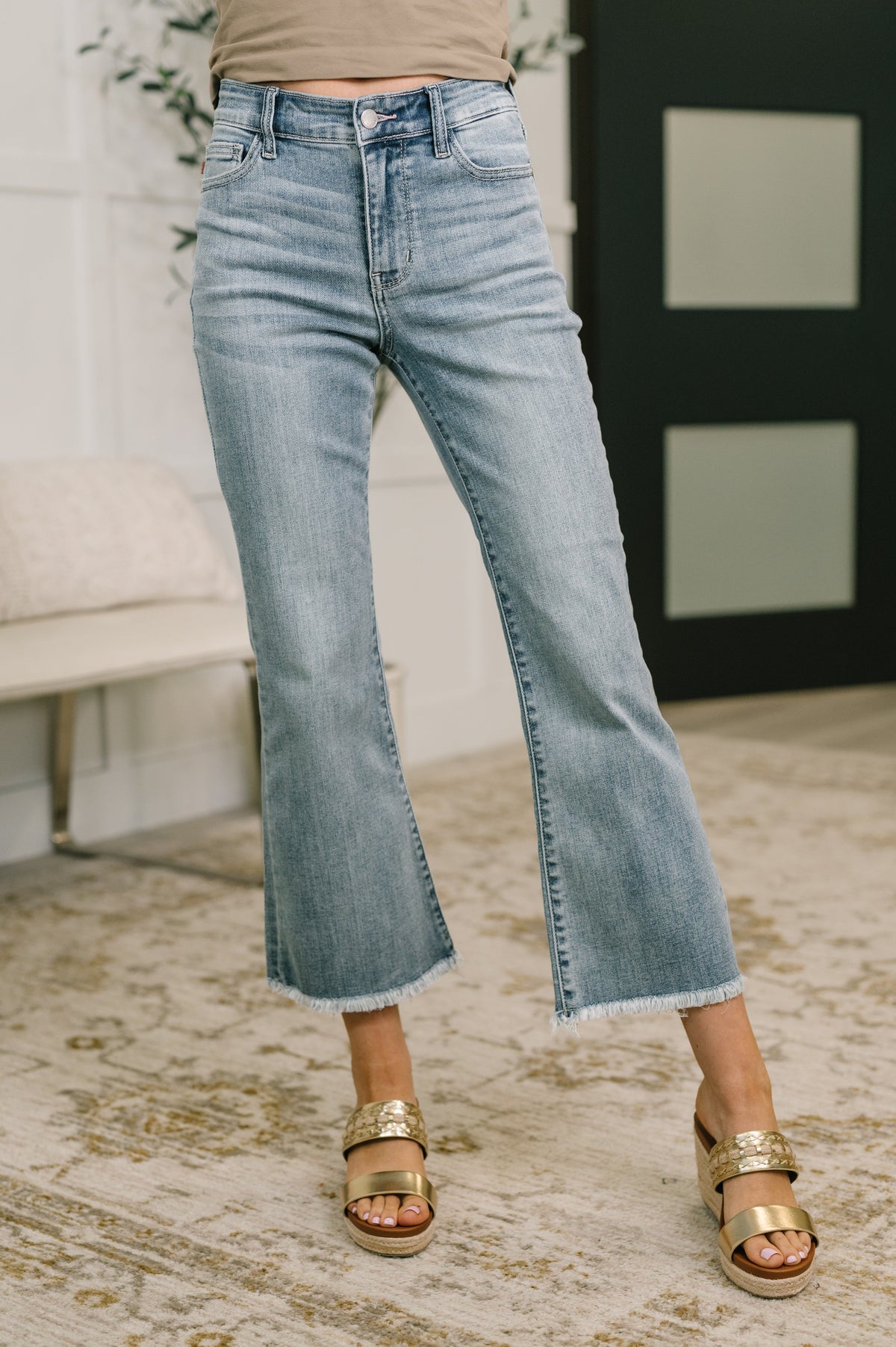 Person wearing light blue jeans and gold sandals in a room with a white wall and plant.