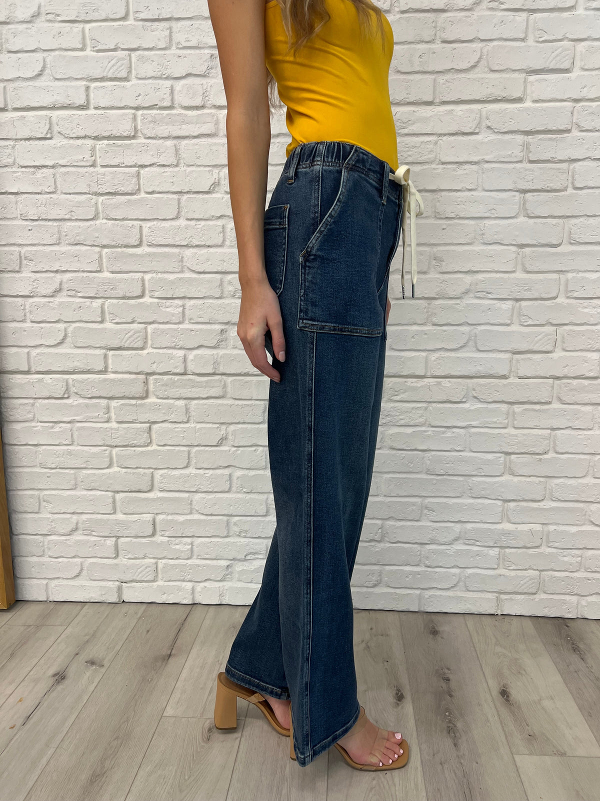 Person wearing blue jeans and a yellow top against a white brick wall.