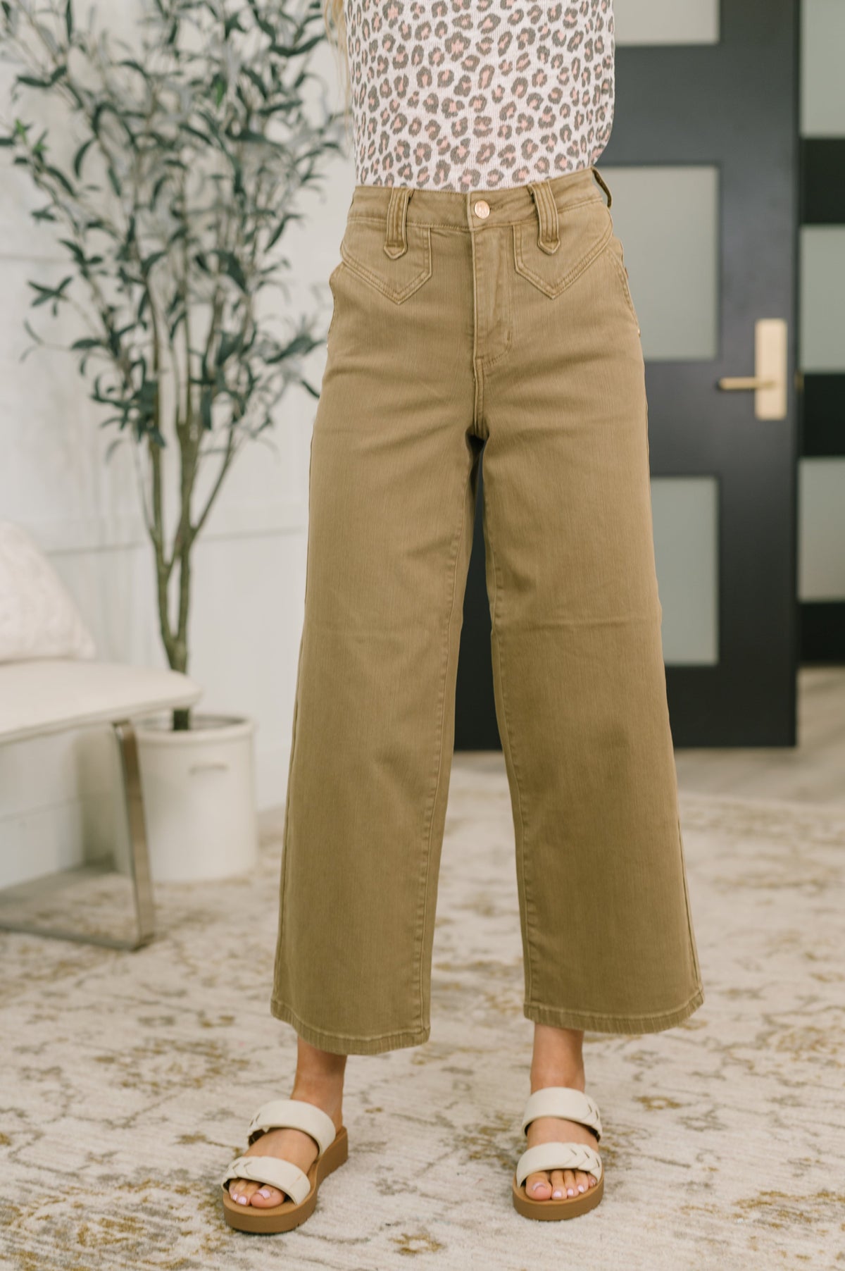 Person wearing tan wide-leg pants in an indoor setting with a plant and door in the background.