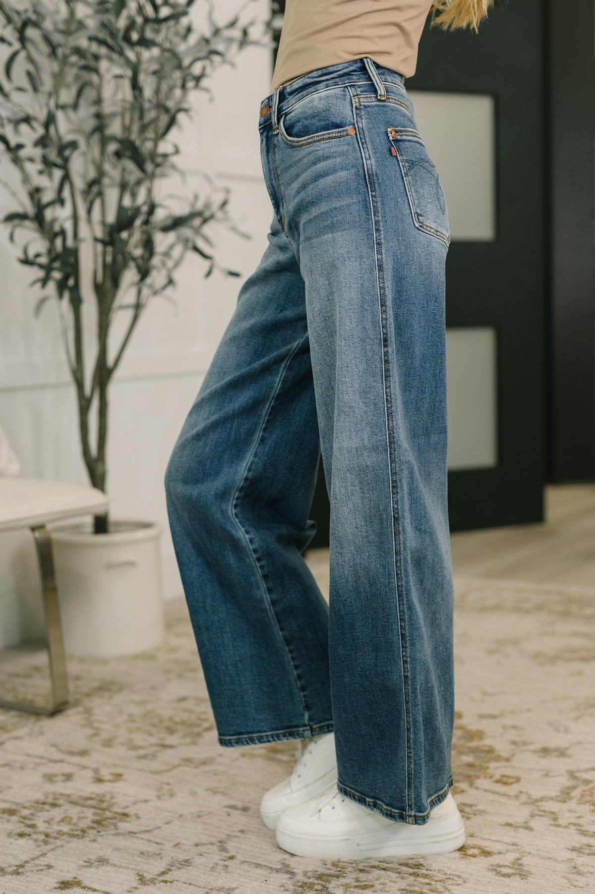 Person wearing blue jeans and white sneakers in a room with a plant and furniture.