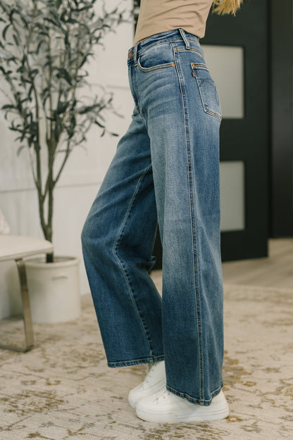 Person wearing blue jeans and white sneakers in a room with a plant and furniture.