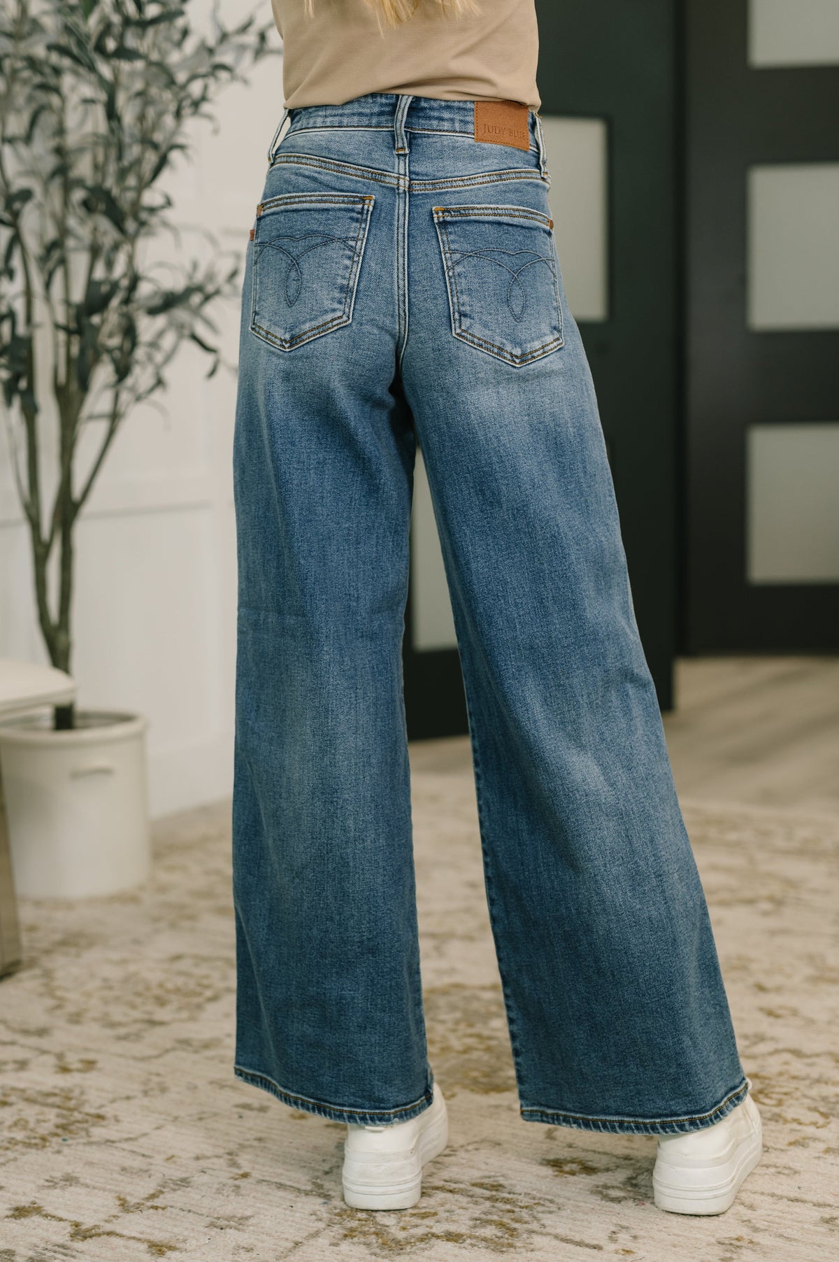 Person wearing blue jeans standing indoors with a plant and wall in the background