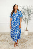 Woman wearing a blue and white patterned dress standing in a room with a plant and tiled floor.