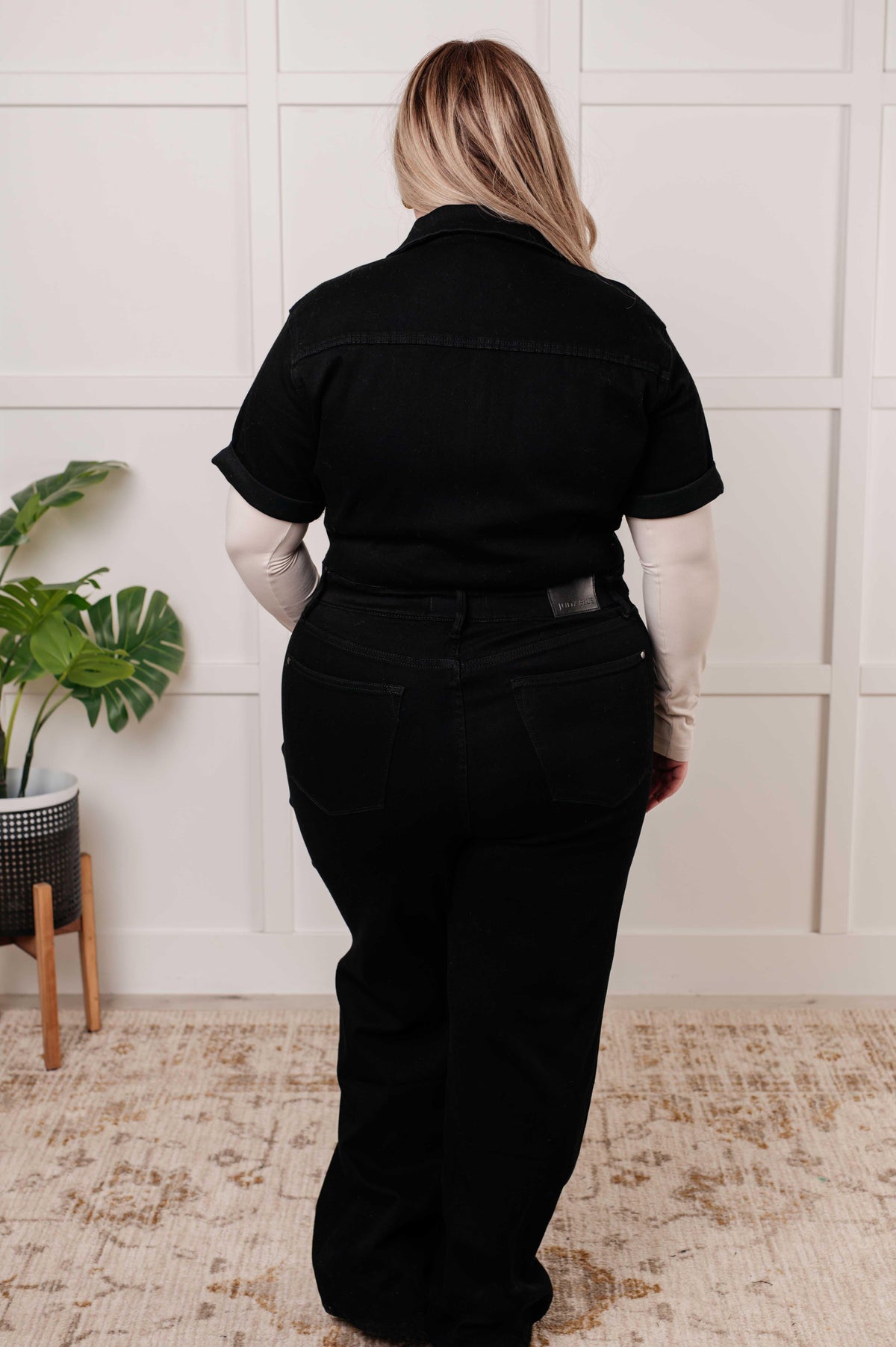 Person wearing a black jumpsuit outfit standing in a room with a plant and white wall.