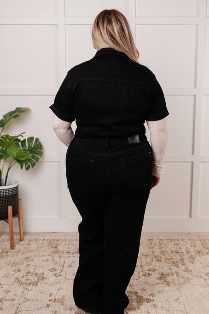 Person wearing a black jumpsuit outfit standing in a room with a plant and white wall.
