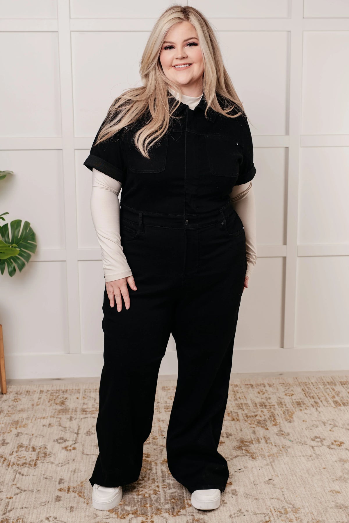 Person wearing a black one-piece outfit with a white shirt underneath, standing against a light-colored wall.