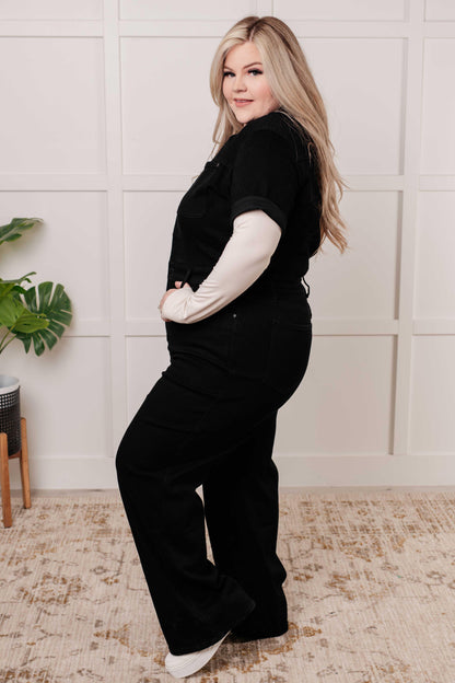 Woman wearing a black one-piece outfit with white sleeves in a room with a plant and rug.