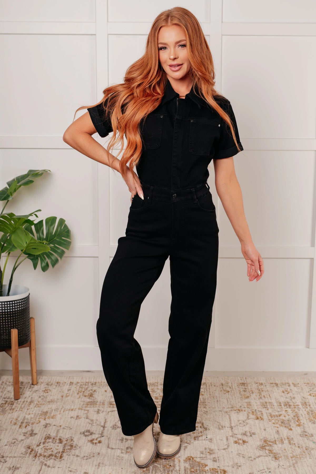 Woman wearing a black one-piece outfit standing in a room with a plant and white wall.