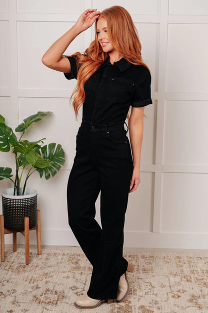 Woman wearing a black one-piece outfit standing indoors with a plant in the background
