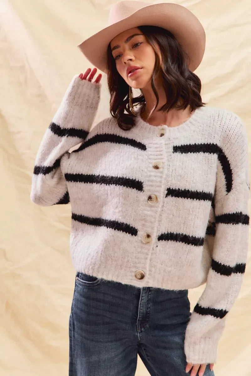 Woman in a cozy striped button-up cardigan, jeans, and a light tan hat, neutral background.