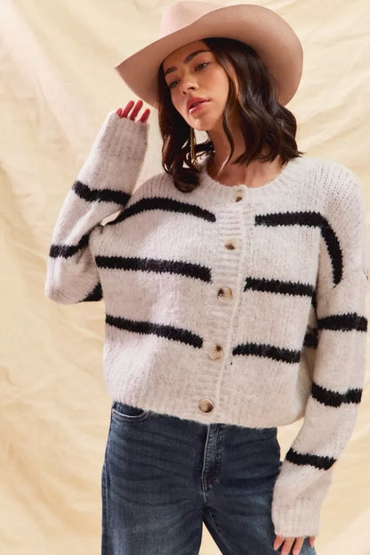 Woman in a cozy striped button-up cardigan, jeans, and a light tan hat, neutral background.