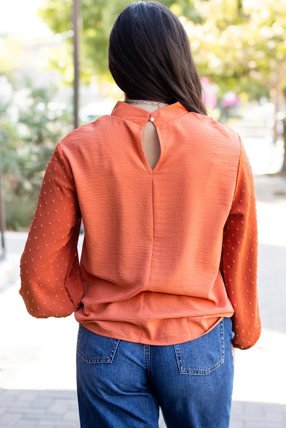 Woman wearing textured orange long-sleeve blouse with keyhole back and blue jeans outdoors