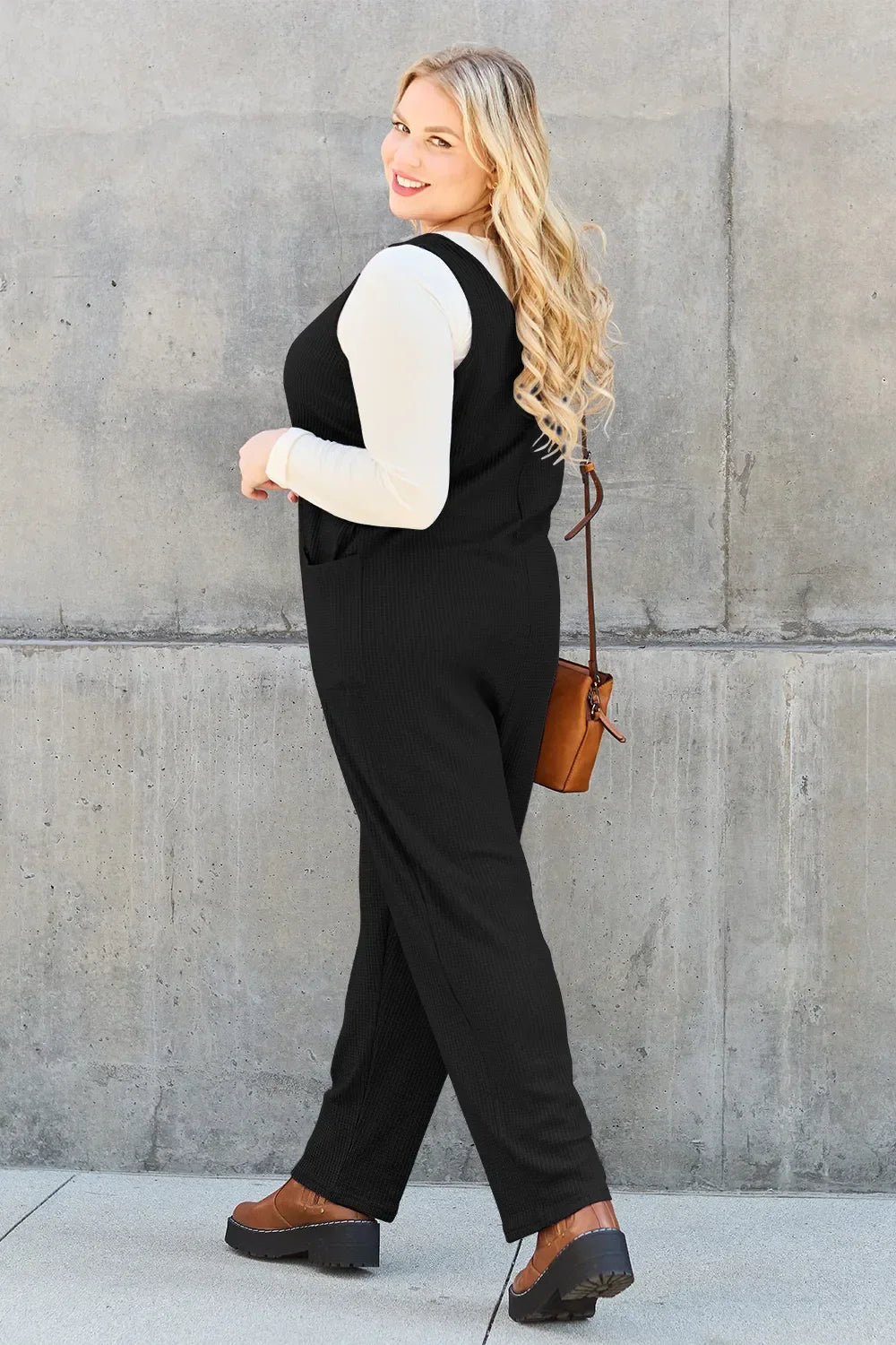 Woman in black sleeveless jumpsuit, cream long sleeve top, brown crossbody bag, brown shoes, standing outdoors