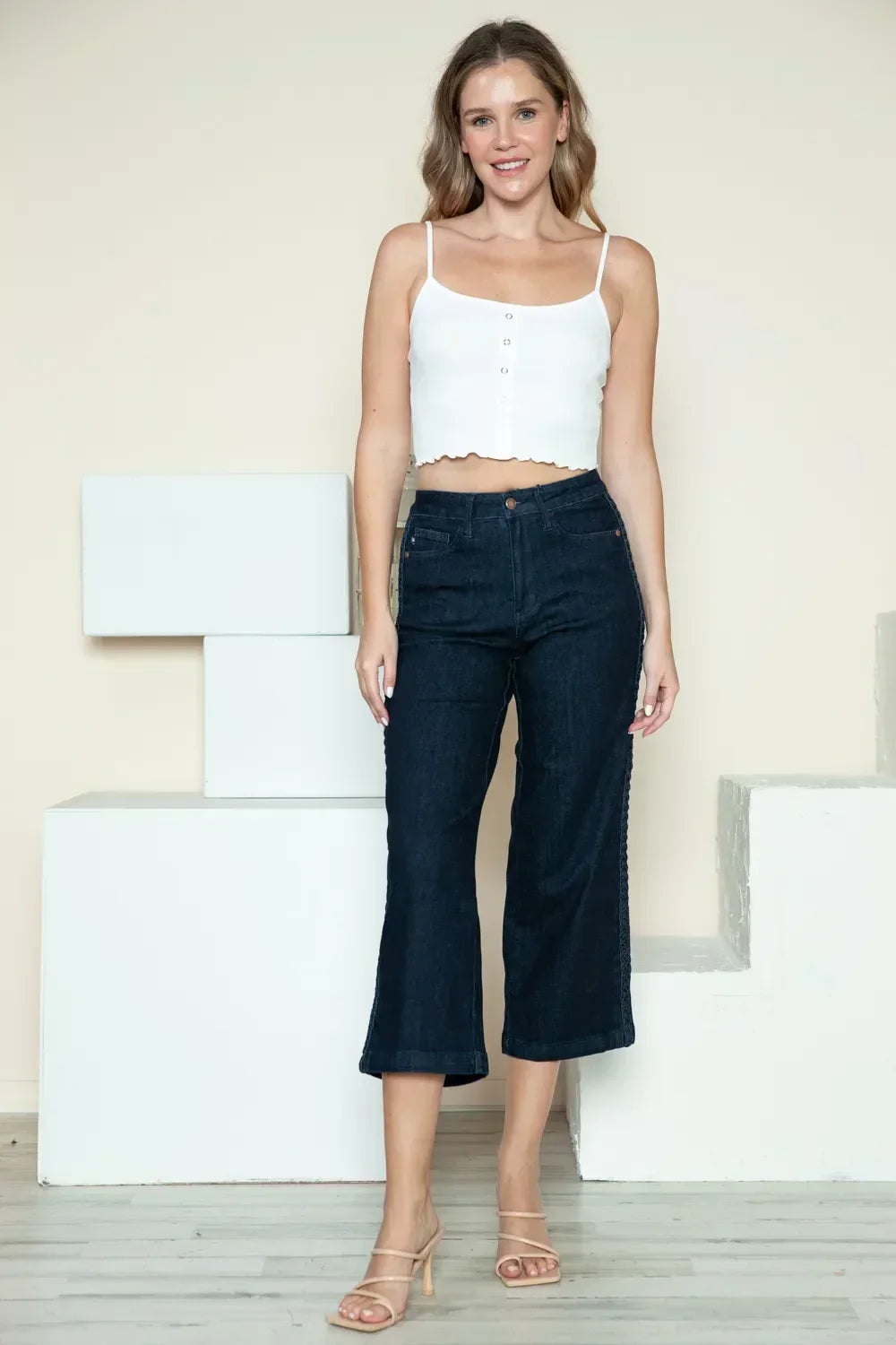 Woman wearing white cropped tank top and dark wide-leg jeans, neutral indoor backdrop