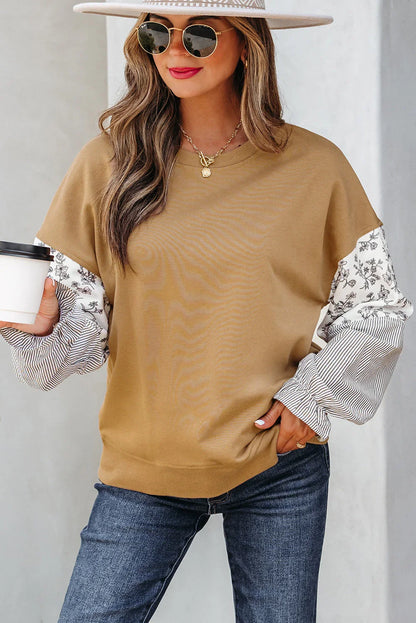 Woman wearing a tan pullover with floral and striped sleeves, blue jeans, wide-brim hat, and sunglasses