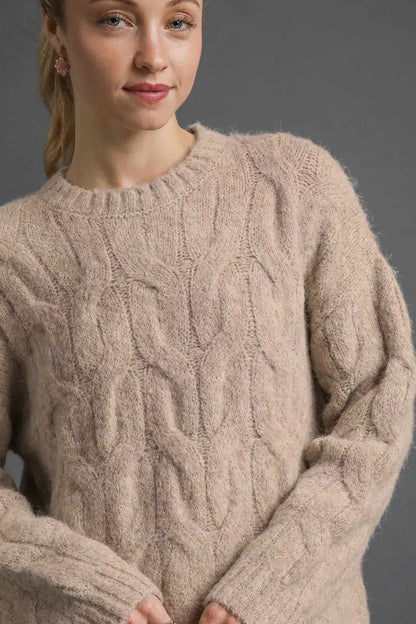 Woman wearing a textured cable knit sweater in light stone color against a gray background.