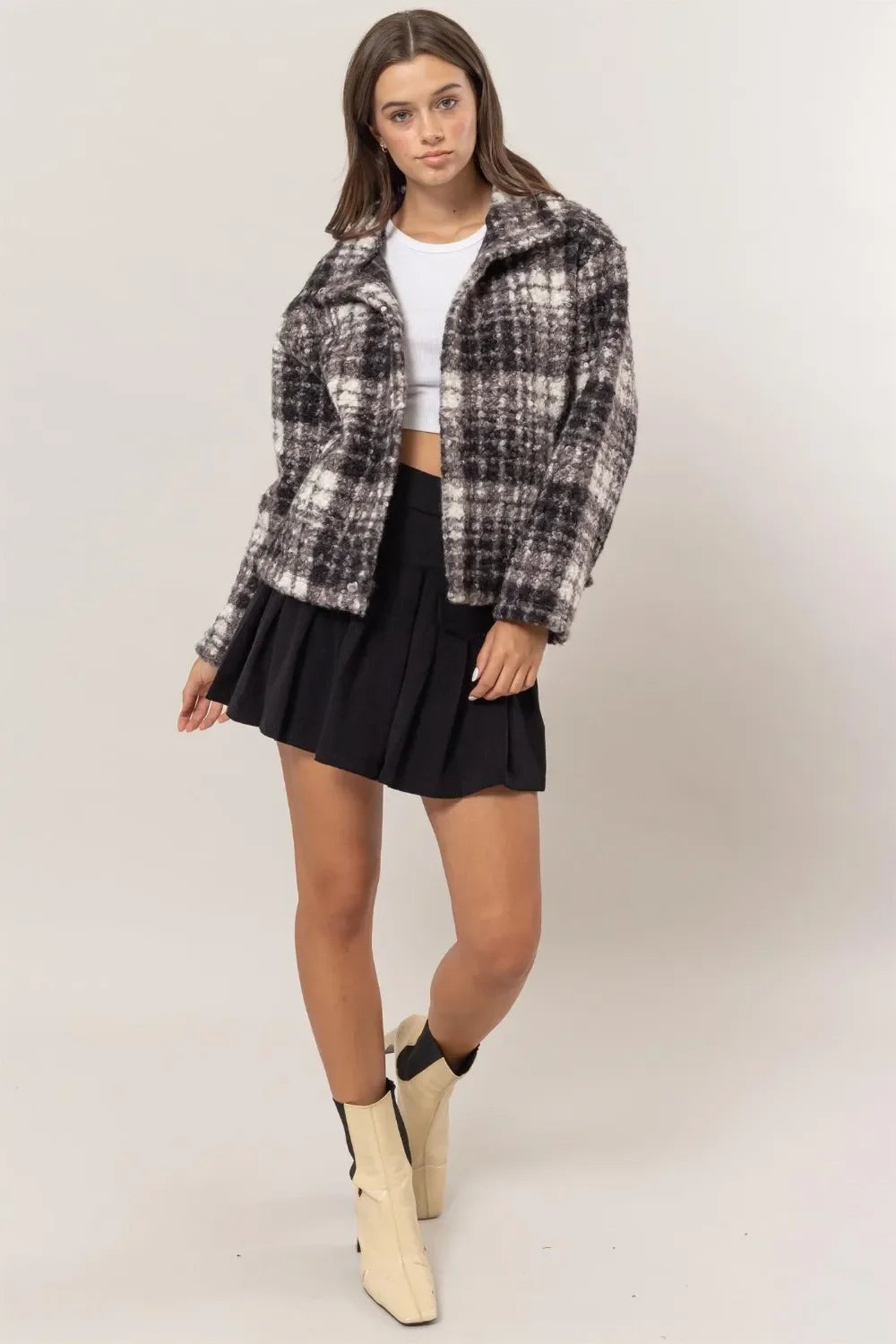 Woman wearing a plaid jacket, white crop top, black pleated skirt, and cream ankle boots