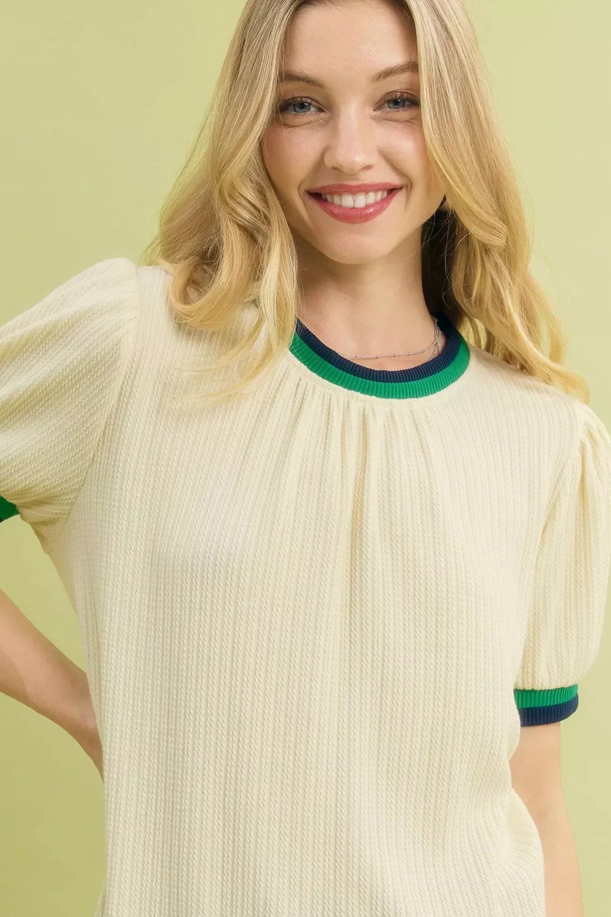 Woman wearing an ecru ribbed knit top with green and navy trim, short puff sleeves, yellow background.