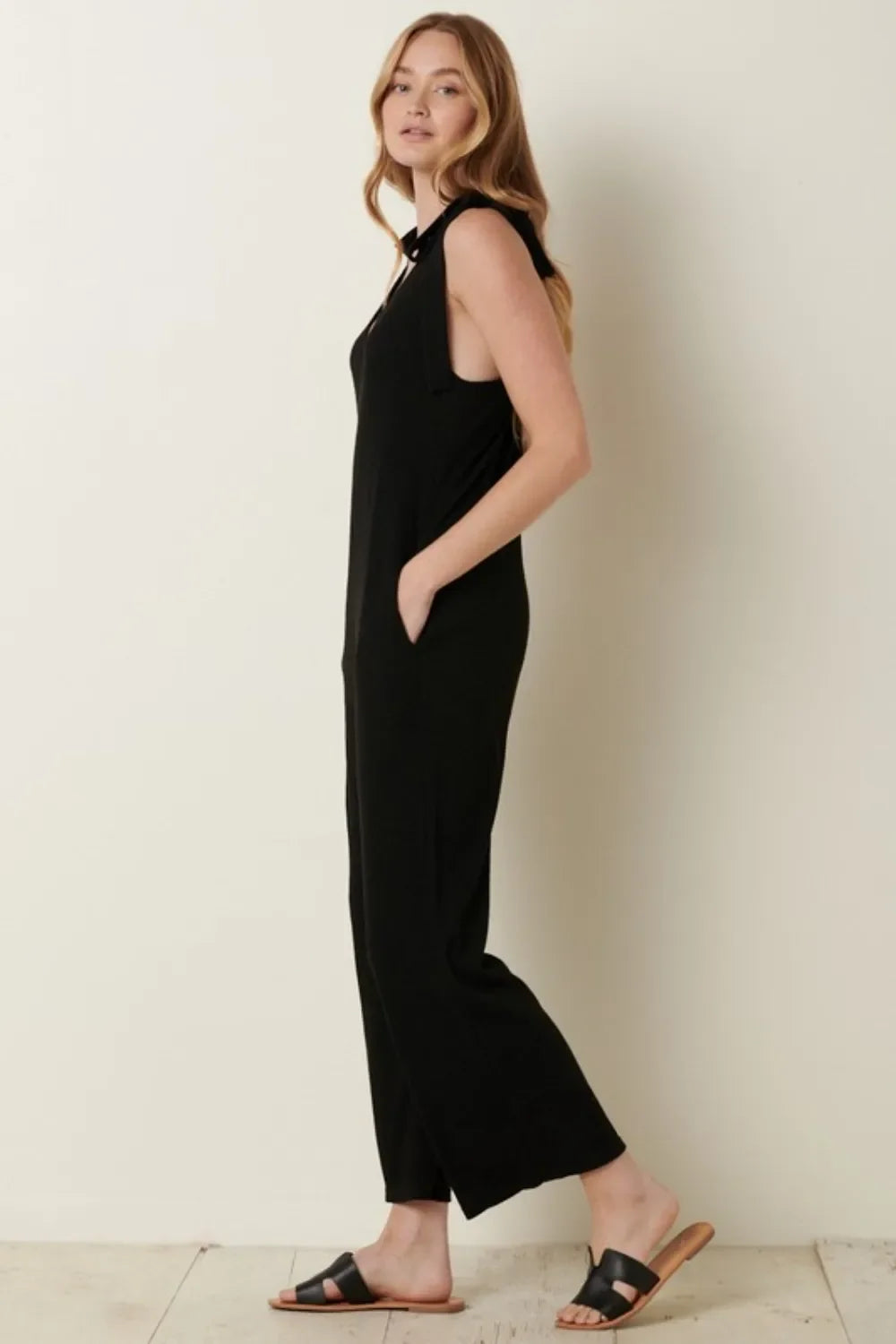 Woman in sleeveless black jumpsuit with side pockets and black sandals, standing indoors